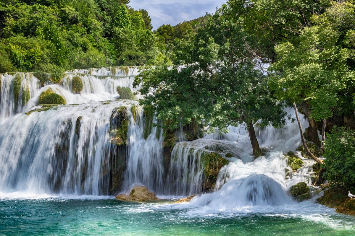A wide multi-tiered waterfall flows into a turquoise pool, surrounded by thick green trees and mossy rocks.