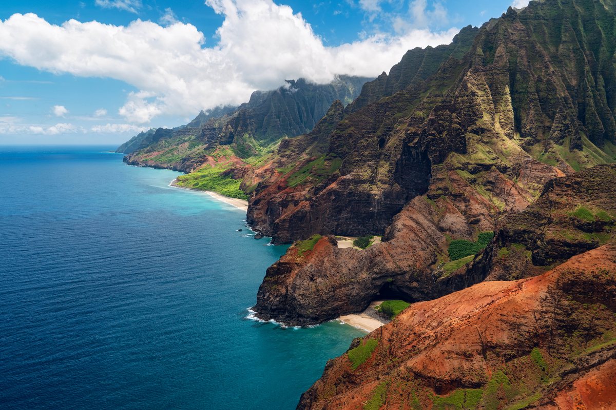 An aerial view of steep, rugged, multi-colored cliffs meeting a deep blue ocean, with small sandy beaches tucked into the base of the rock faces.