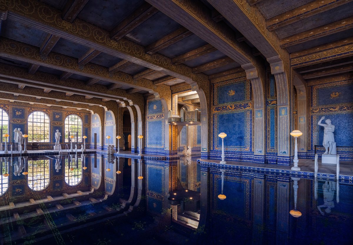 An ornate indoor swimming pool room featuring deep blue tile walls with gold trim, arched windows, decorative ceiling beams, and white statues standing on pedestals along the water's edge, with the entire scene reflected in the still pool water.