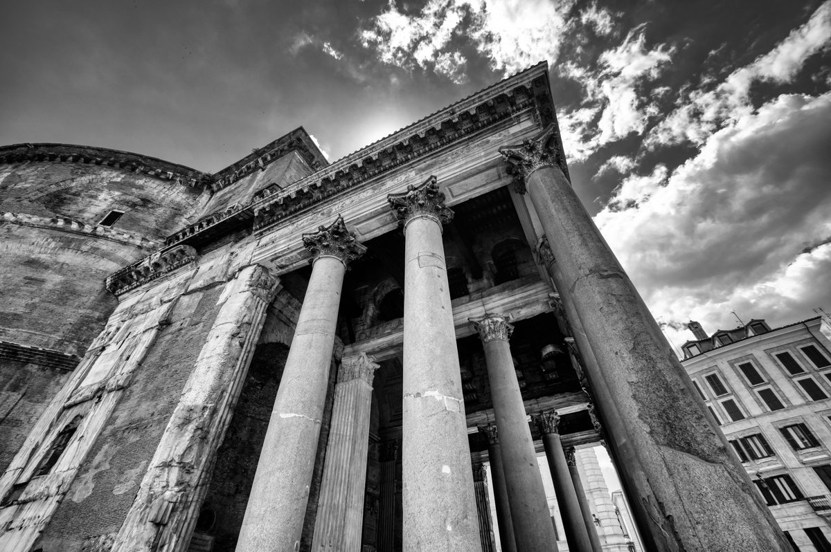 A low-angle black and white photograph of a classical stone building with large Corinthian columns under a cloudy sky.