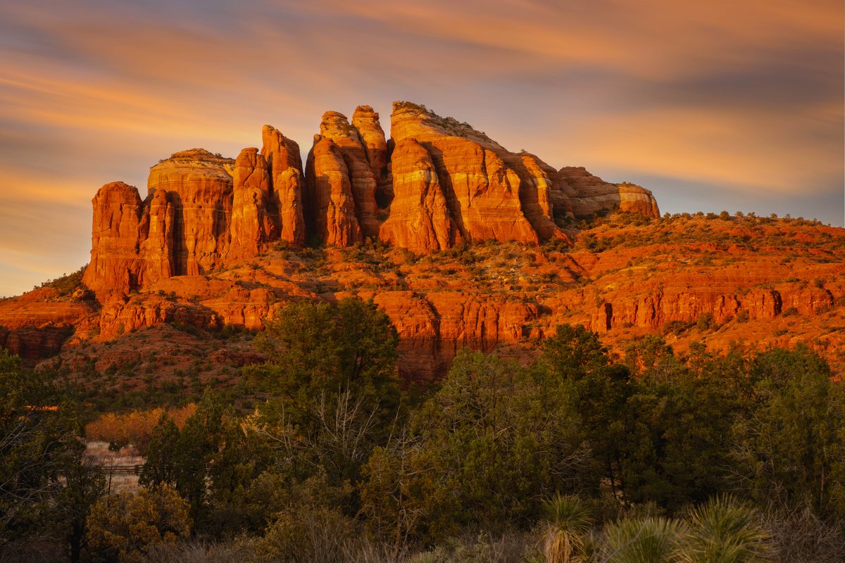 A large, jagged red rock mountain formation illuminated by warm sunset light, with trees and shrubs in the foreground under a streaked orange sky.