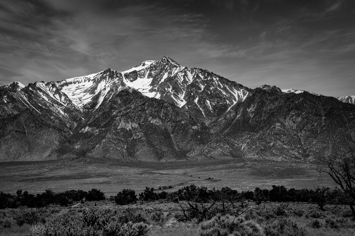 A black and white landscape showing a tall, snow-capped mountain peak looming over a vast, arid valley floor with sparse desert vegetation.