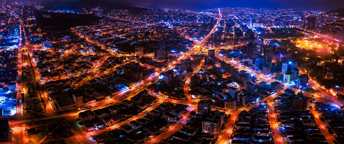 An aerial nighttime view of a densely populated city with a network of orange-lit streets and various building clusters illuminated against a dark background.
